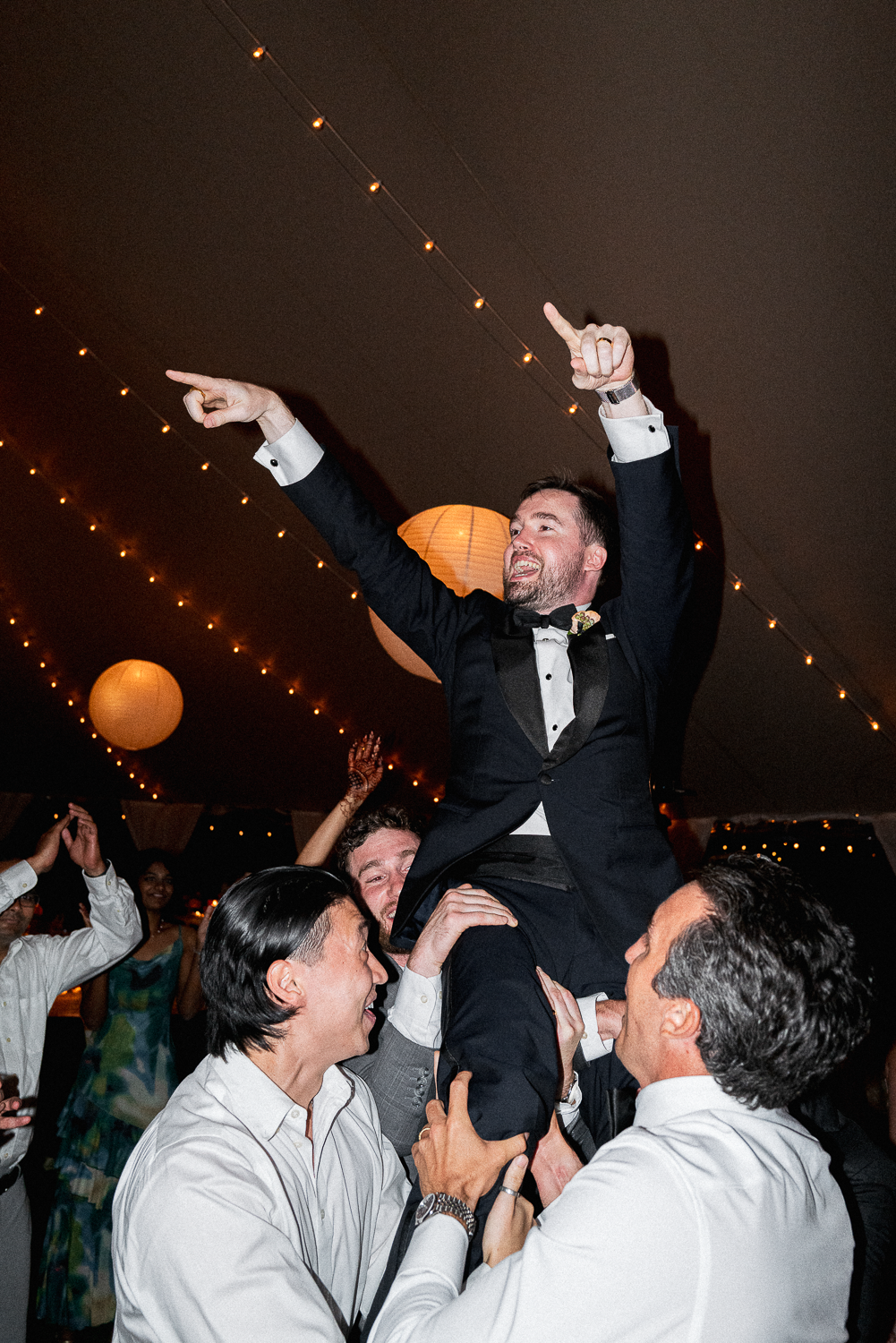 A man dressed in a tuxedo being lifted and celebrated by others at a wedding reception under string lights and paper lanterns.