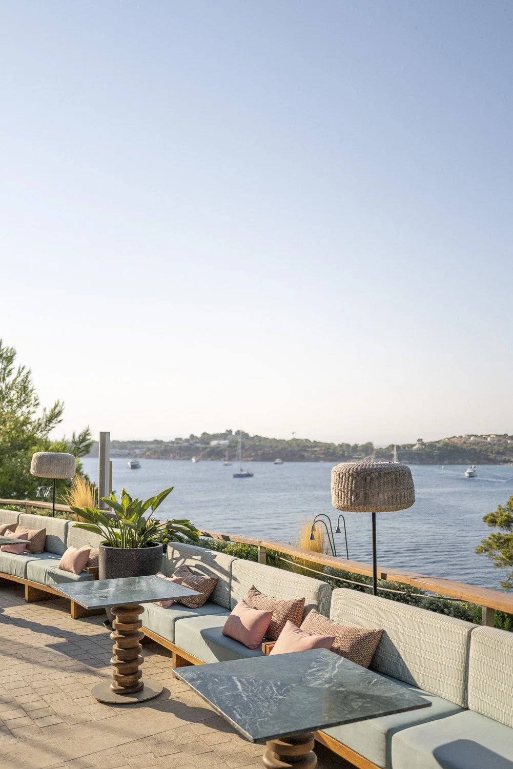 Outdoor patio with seating area, pink pillows, large potted plant, and view of a bay with sailboats and hilly landscape in the background.