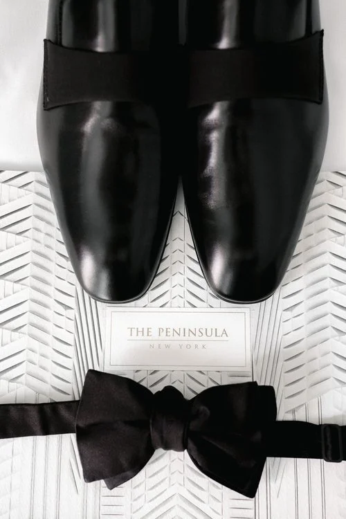 Pair of polished black shoes with elastic strap, a black bow tie, and a gift box from The Peninsula New York.