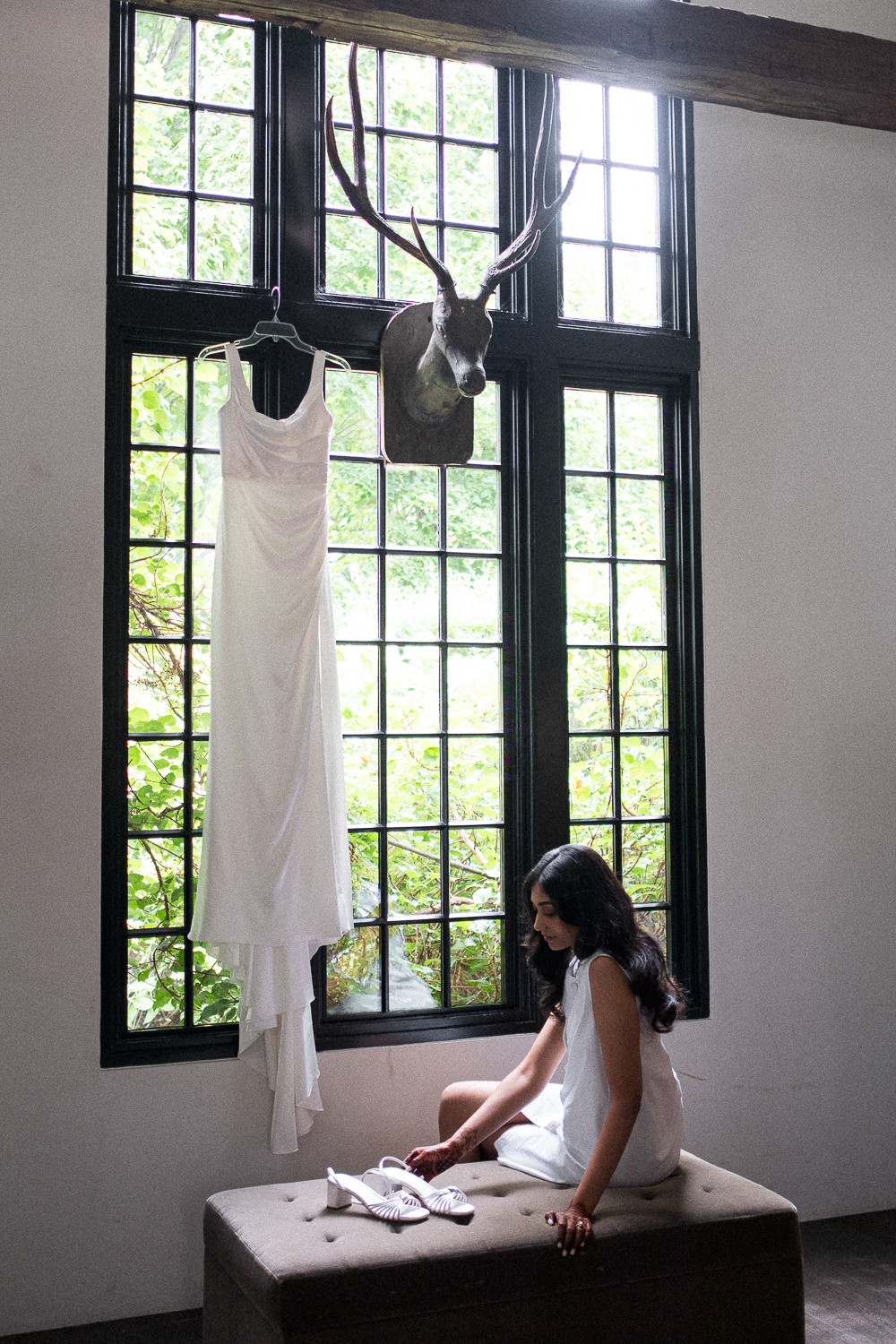 A woman in a white dress sitting on a bench by a large window, with a white dress hanging on the window frame and a deer head mounted on the wall above.