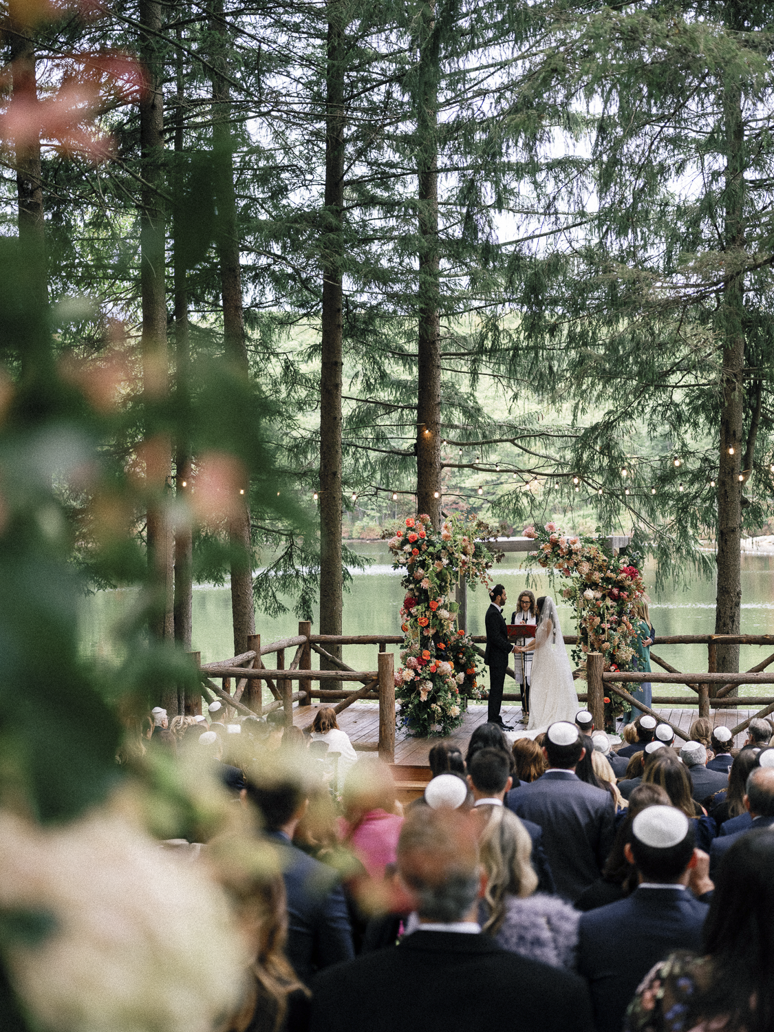 A wedding ceremony takes place outdoors by a lake, with a decorated floral arch and string lights amidst tall trees, and guests in formal attire watching.