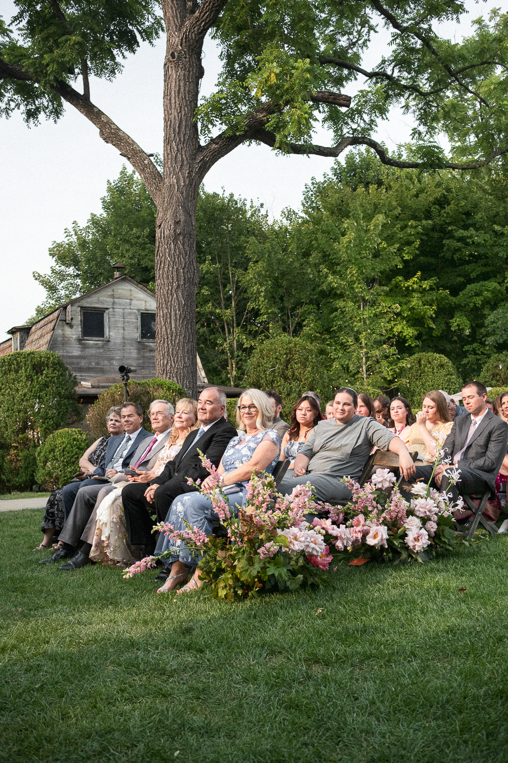 People seated outdoors during a formal event, with a large tree and greenery in the background, and a flower arrangement in the foreground.