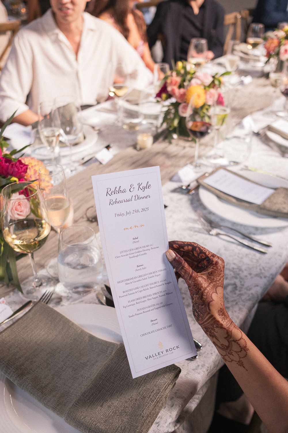 A woman with henna tattoos on her hand holding a menup during a rehearsal dinner, with a decorated table, glasses of wine, and other guests in the background.