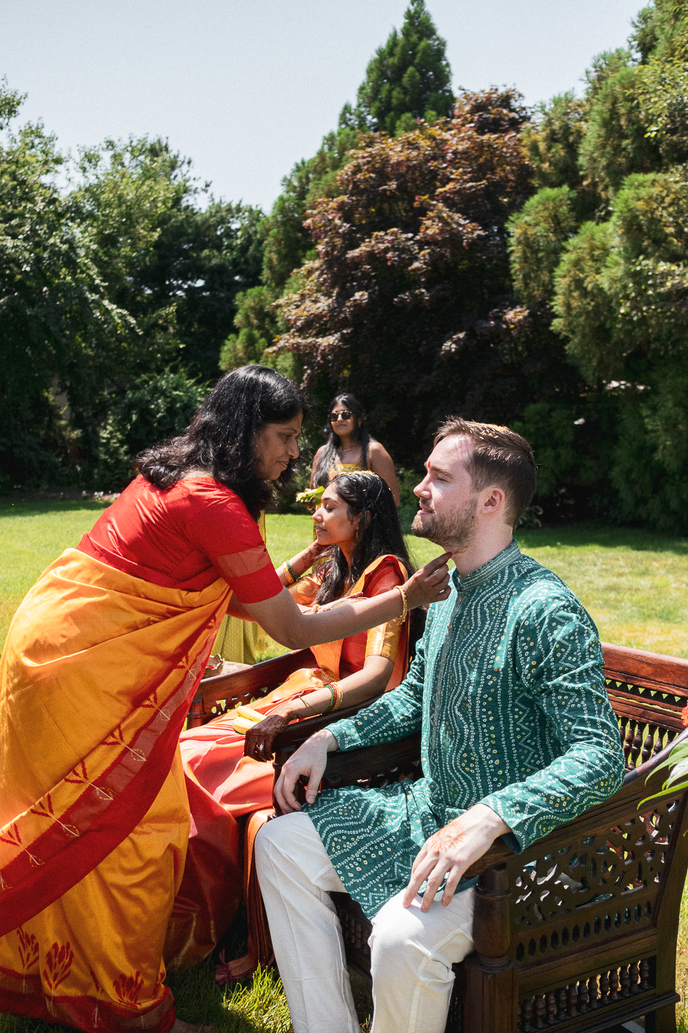 A woman in a red and orange sari applies a mark on the man's forehead, who is seated on a wooden bench, during an outdoor cultural or religious ceremony. Two women in traditional attire are in the background, with lush green trees and grass surroundi