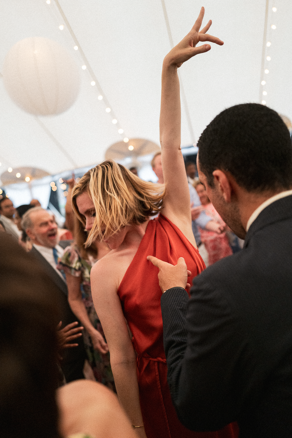 A woman in a red dress dancing with a man at a social event or party under a decorated tent with hanging paper lanterns and string lights.