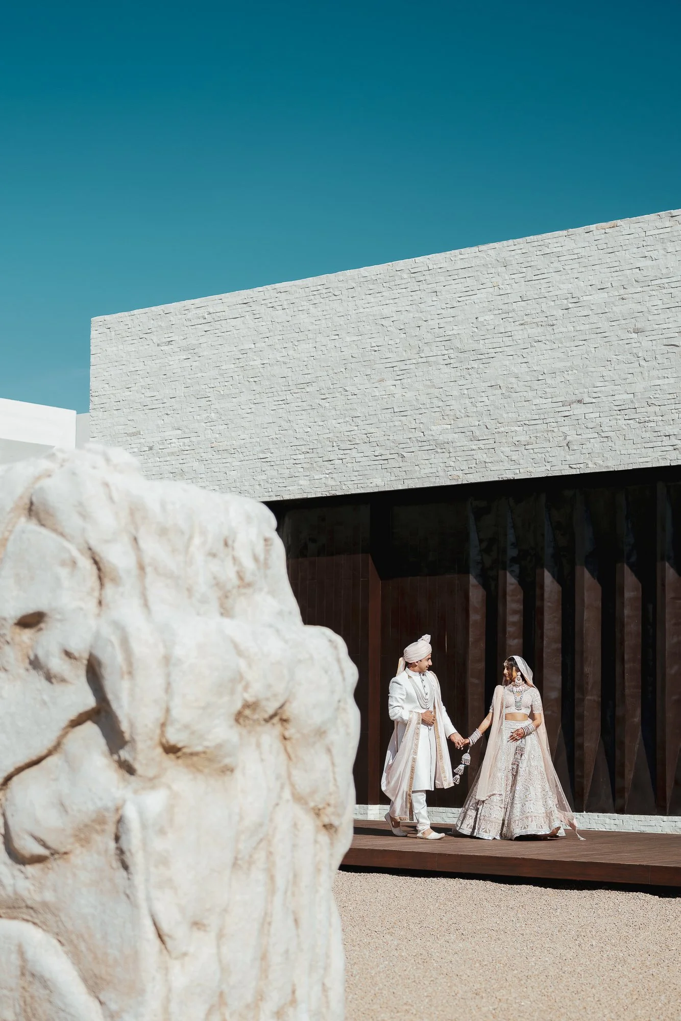 A bride and groom holding hands and walking in traditional Indian wedding attire on a stage, with large white rocks in the foreground and modern architecture in the background.