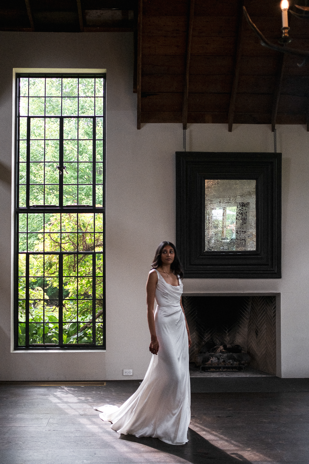 A woman in a white dress stands inside a room with a large window showing green foliage outside, a black framed mirror above a fireplace, and a wooden ceiling.