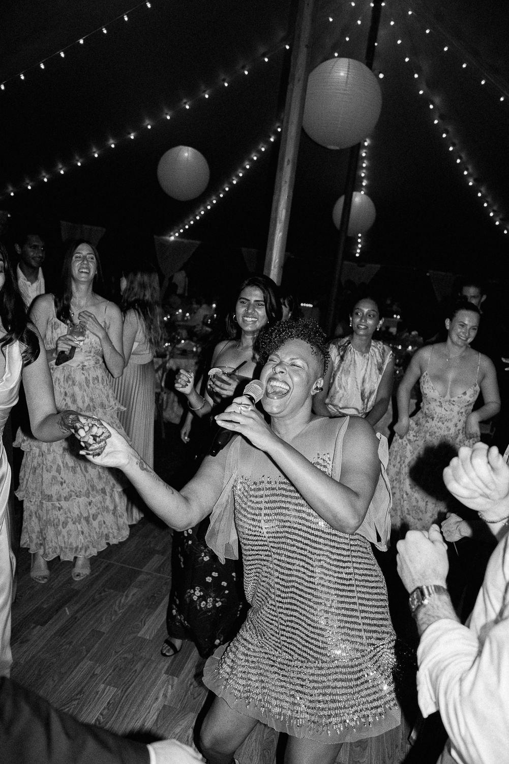 A woman singing into a microphone and dancing at a celebration, surrounded by smiling guests in a decorated tent with hanging paper lanterns and string lights.