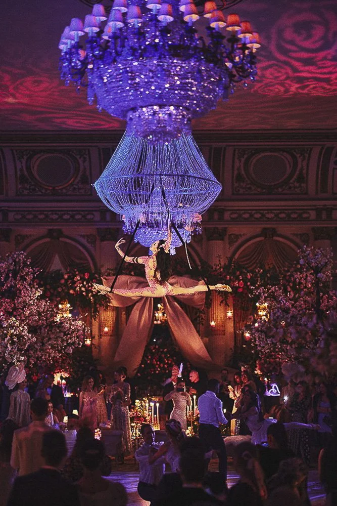 Elegant event hall decorated with a large chandelier, floral arrangements, and a circus or acrobatic theme with a performer in a costume hanging under the chandelier.