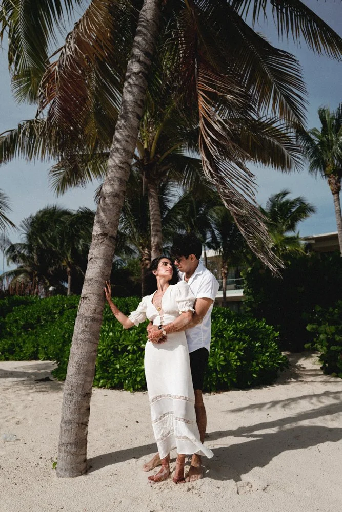 A couple dressed in white on a sandy beach surrounded by palm trees, with the woman leaning against a palm tree and the man standing behind her, holding her waist.