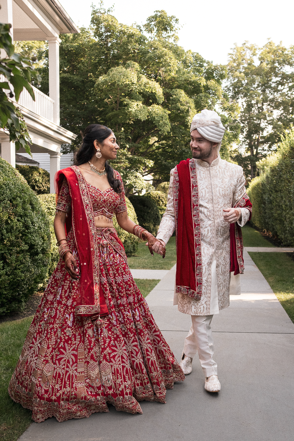 A bride and groom in traditional Indian wedding attire holding hands and smiling at each other outdoors. The bride is wearing a red and gold embroidered lehenga and jewelry. The groom is dressed in a white sherwani with red accents and a turban. They