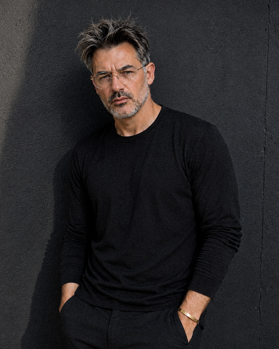 A middle-aged man with salt-and-pepper hair, glasses, and a beard, wearing a black long-sleeve shirt and black pants, leaning against a dark gray wall with his hands in his pockets.