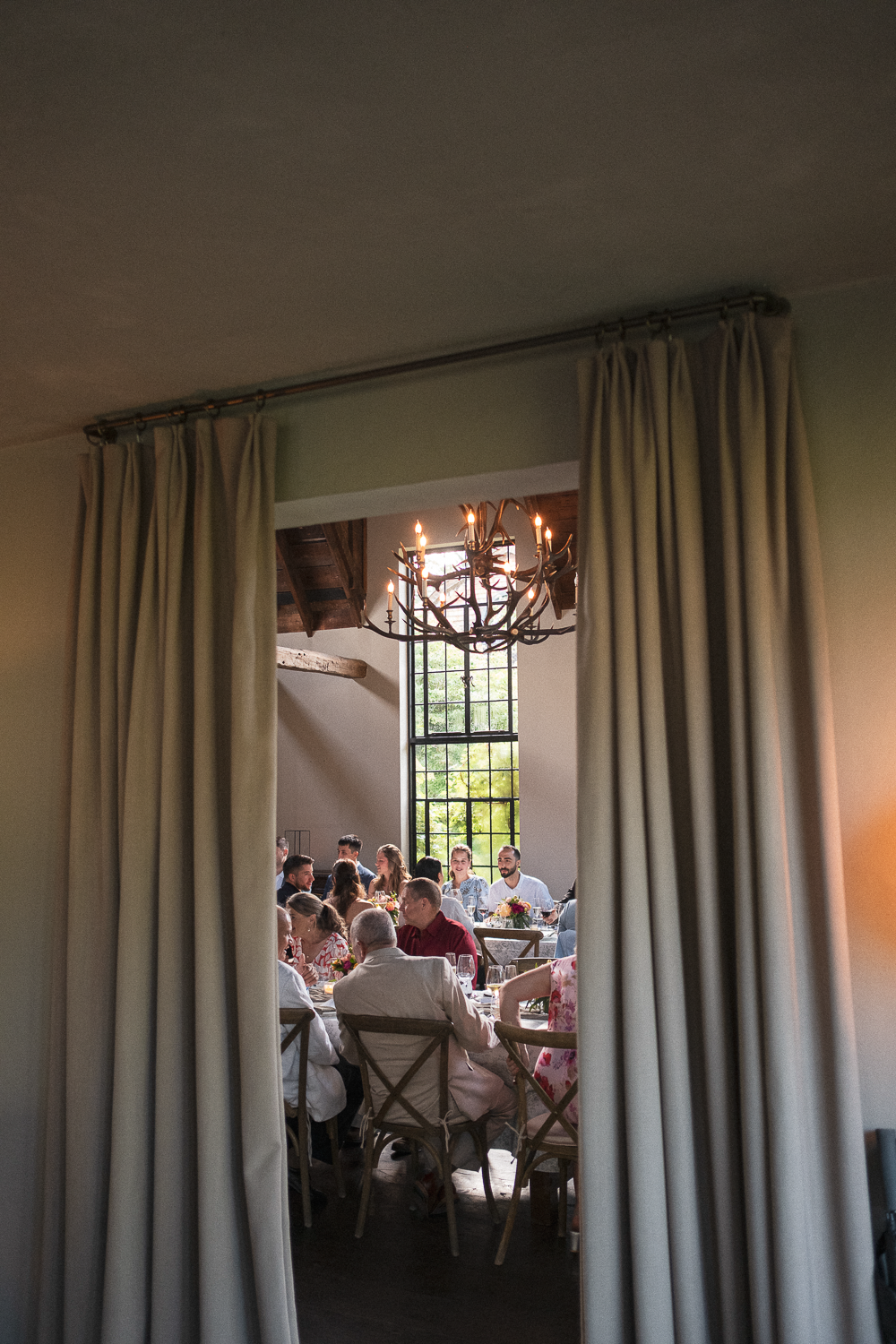 People gathered around a dining table in a well-lit room with large window, chandelier, and wooden ceiling beams, seen through an open doorway with curtains.