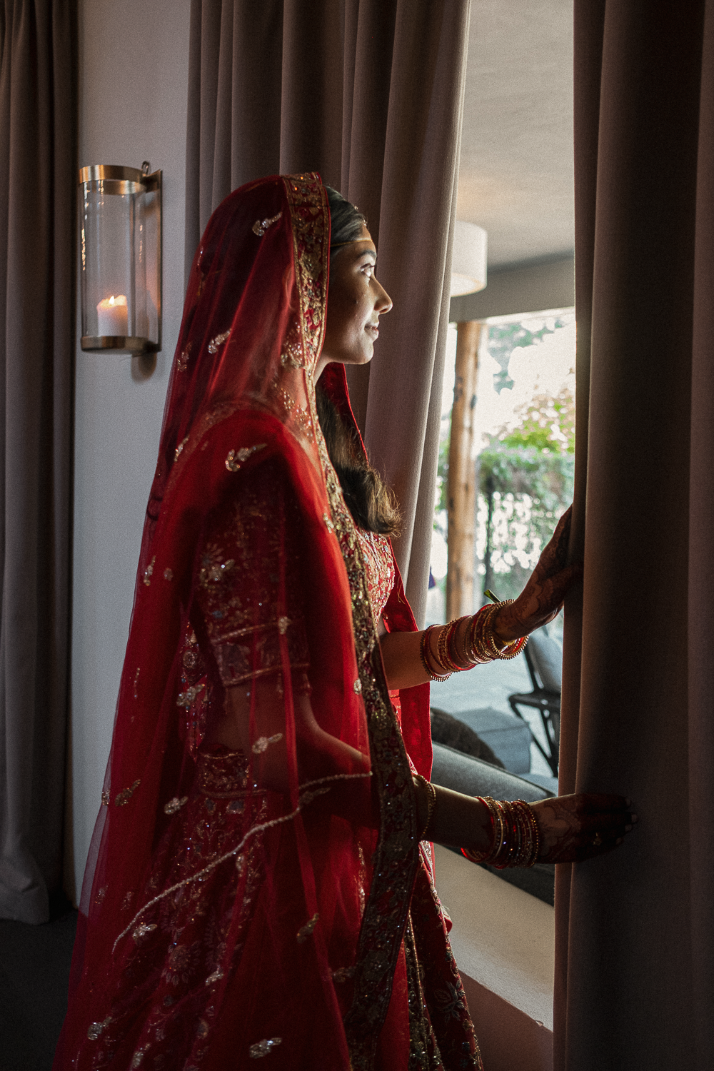 A woman dressed in traditional Indian wedding attire standing by a window, holding back curtains and looking outside.