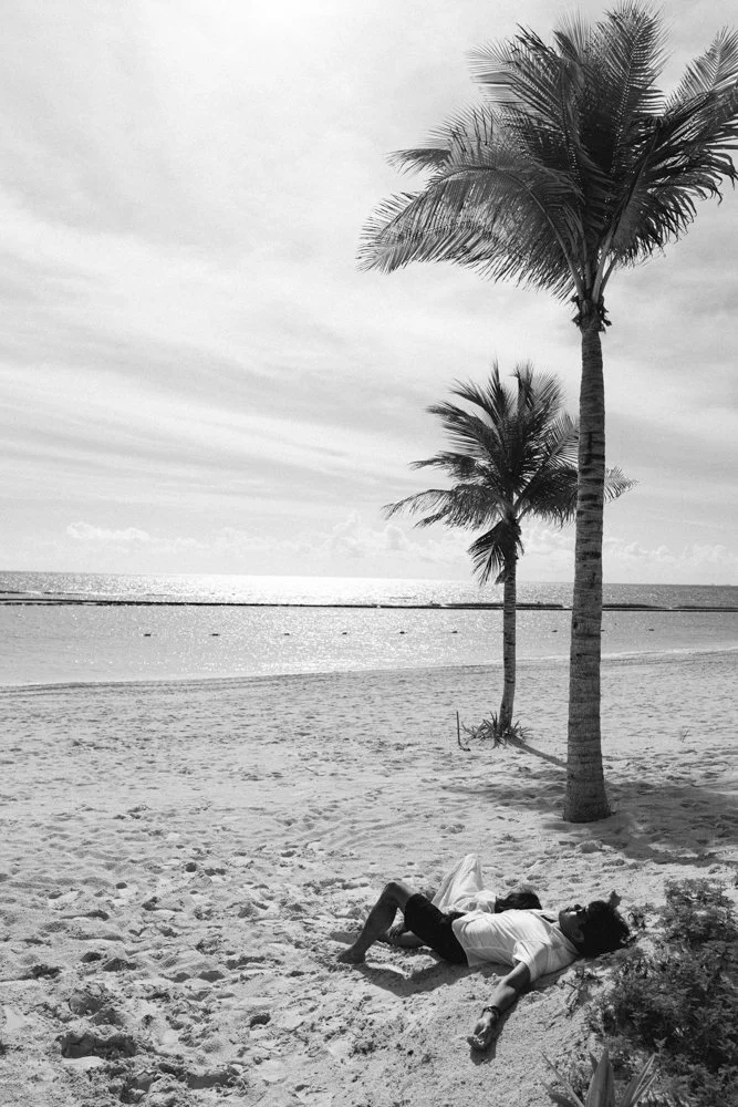 A person lying on the sandy beach under palm trees, with the ocean and sky in the background.