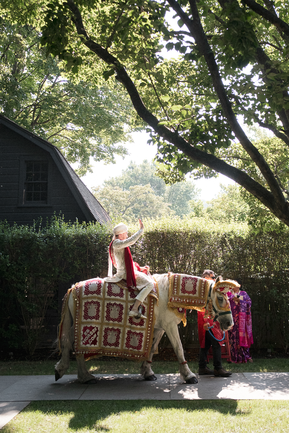 A person dressed in traditional Indian attire riding a decorated white horse, waving, with two people walking beside them in colorful clothing, on a sidewalk with trees and a hedge in the background.