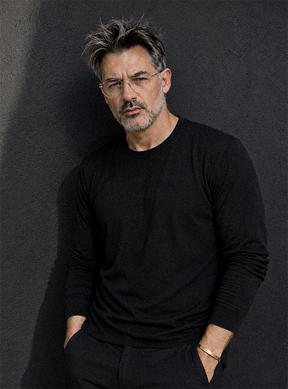 Andreas Sellinidis is standing against a dark wall, wearing a black long-sleeve shirt and dark pants, with a serious expression.