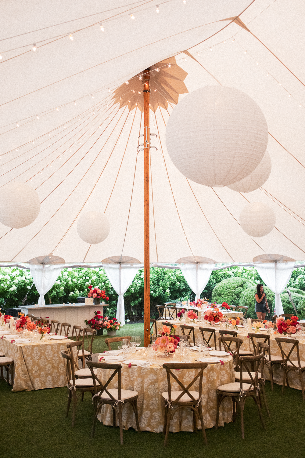 Decorated outdoor event tent with round tables, floral centerpieces, string lights, and a woman standing near the back.