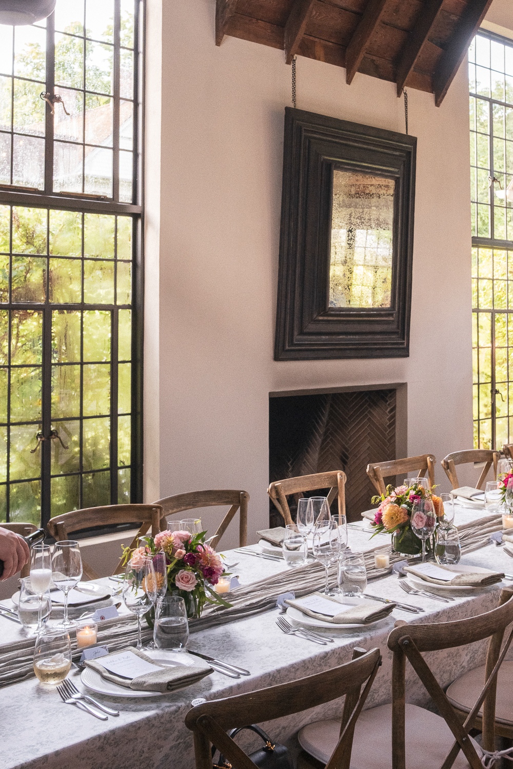 A table set for a formal meal with floral centerpieces, candles, wine glasses, and place settings, inside a room with large windows and a fireplace.