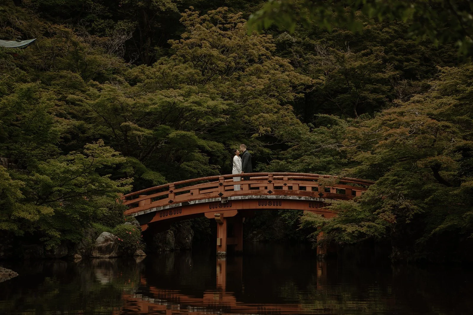 Couple standing on an arched vermilion bridge over a reflecting pond in a Kyoto garden