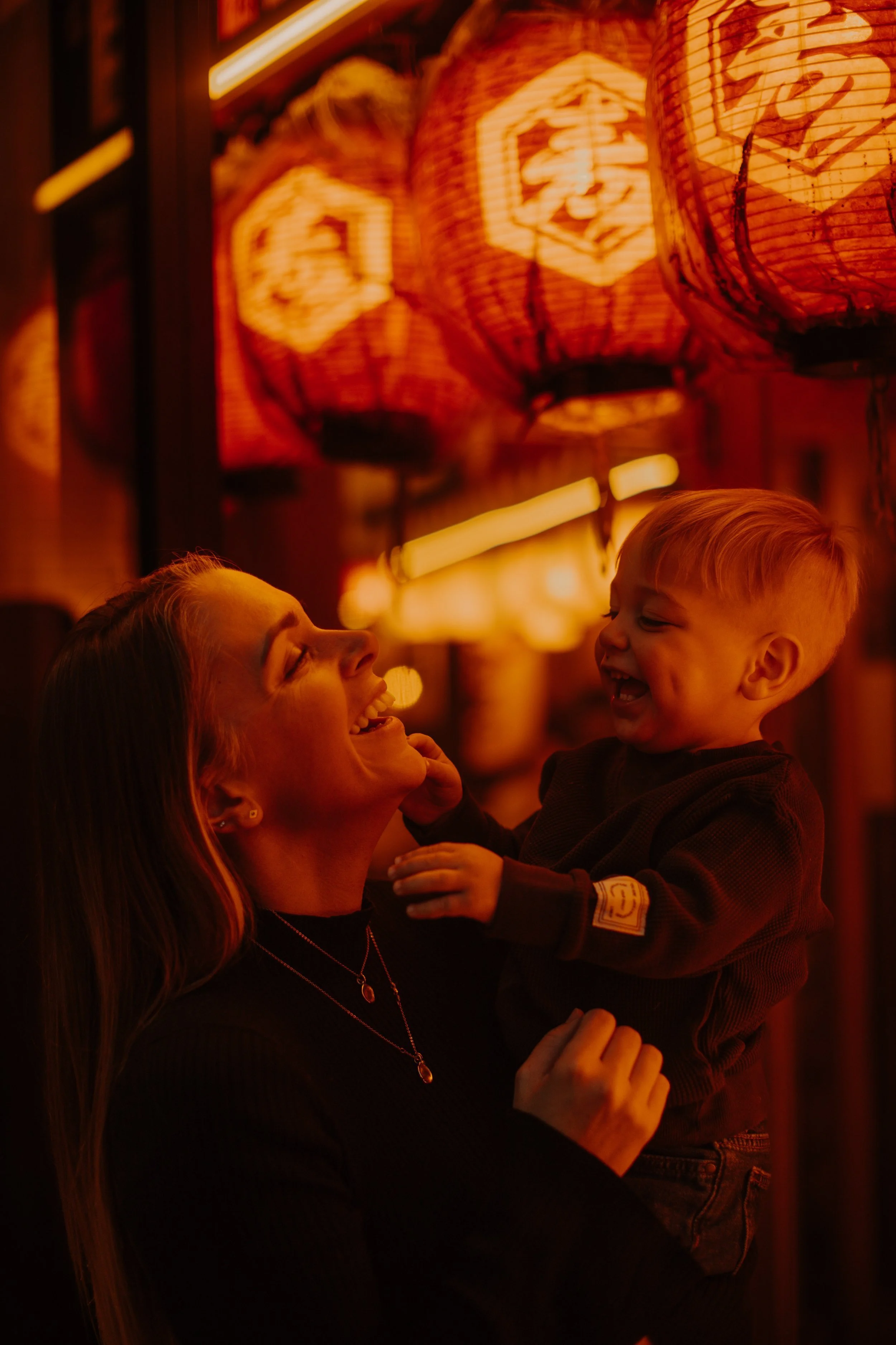 Mother and toddler son laughing together beneath red paper lanterns on a Tokyo backstreet.