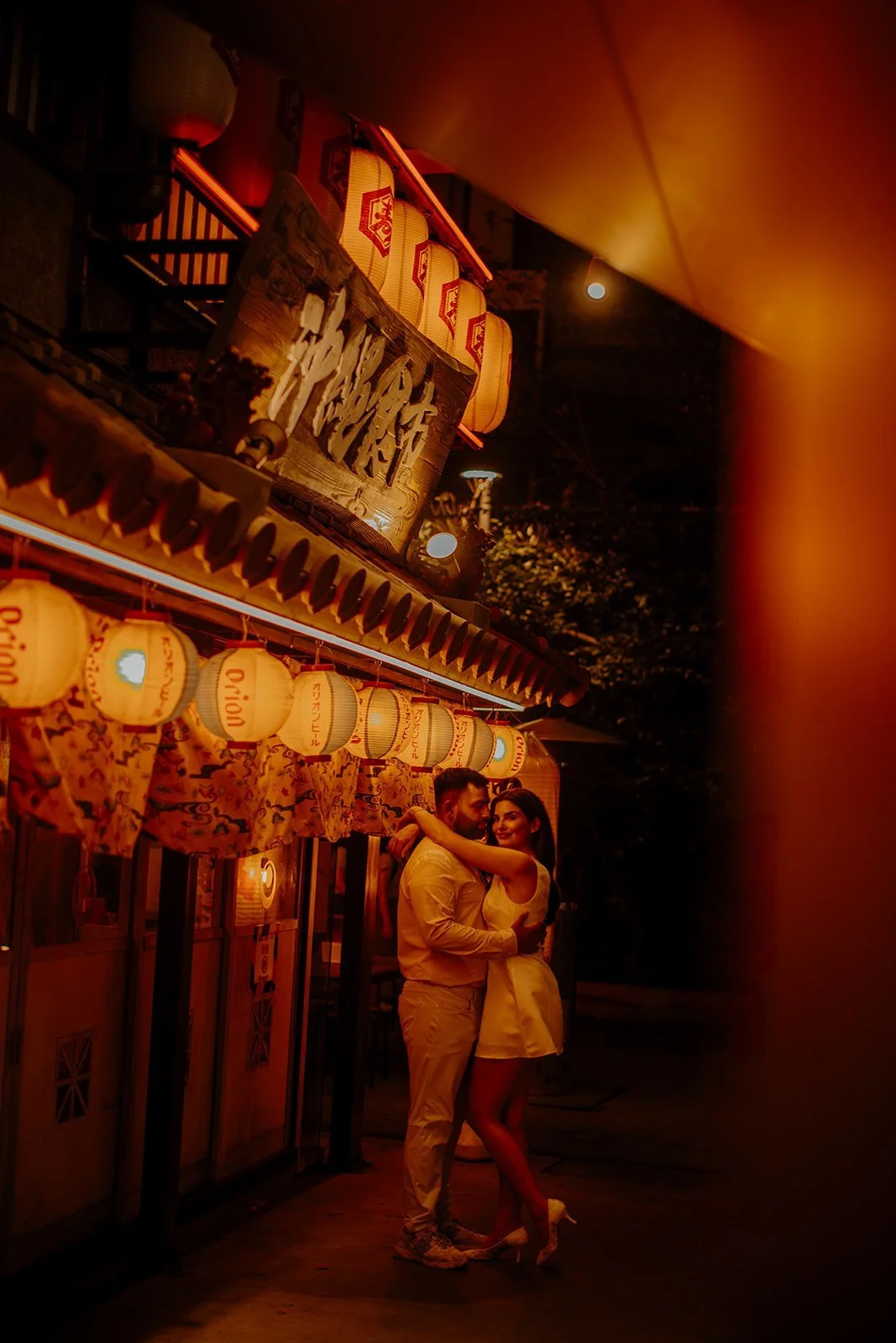 Couple embracing beneath red lanterns outside an Okinawan food stall in a Tokyo yokocho alley