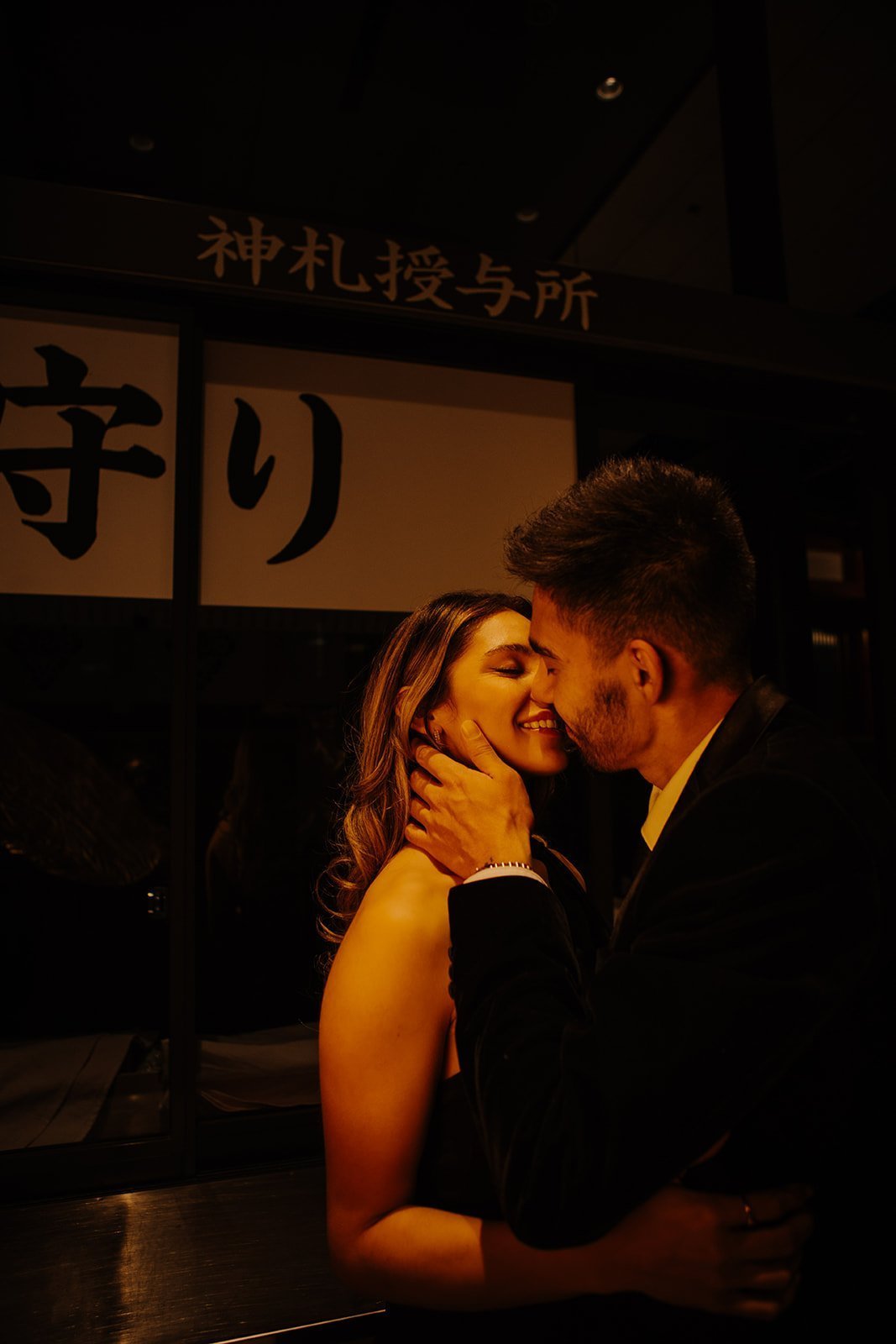 Couple sharing a kiss beside a lantern-lit shrine amulet counter during a nighttime engagement session in Tokyo.