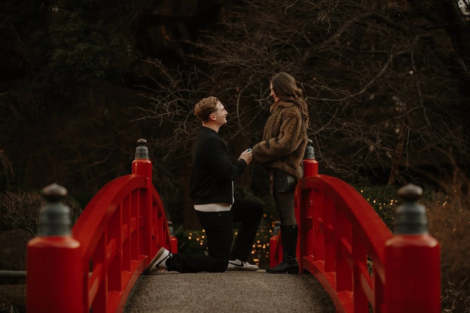 Newly engaged couple smiling on a red bridge moments after a surprise proposal in a Tokyo garden.

