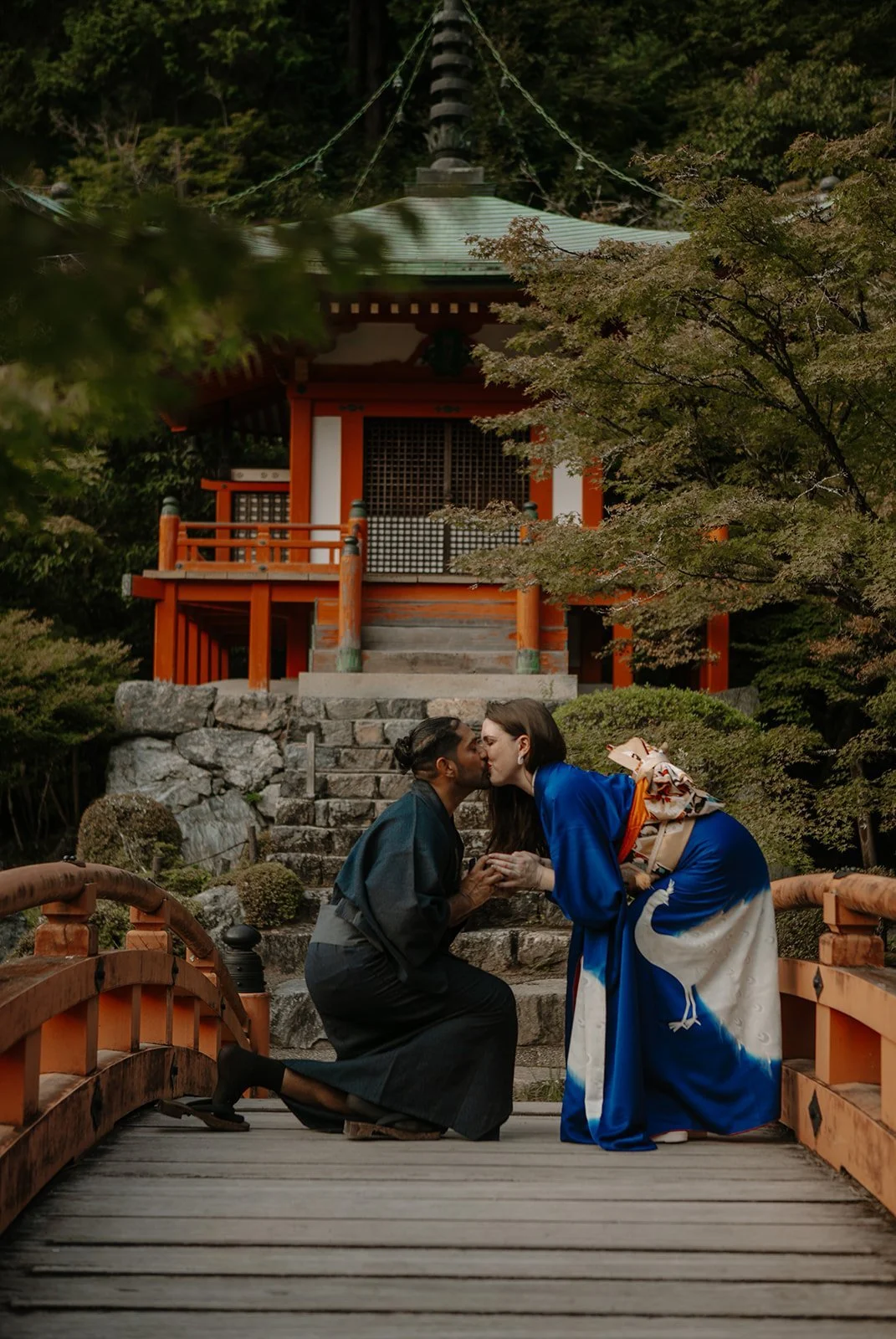 Man kneeling to propose to his partner, dressed in traditional kimono, kissing on an orange bridge in front of a small shrine pavilion in Japan.