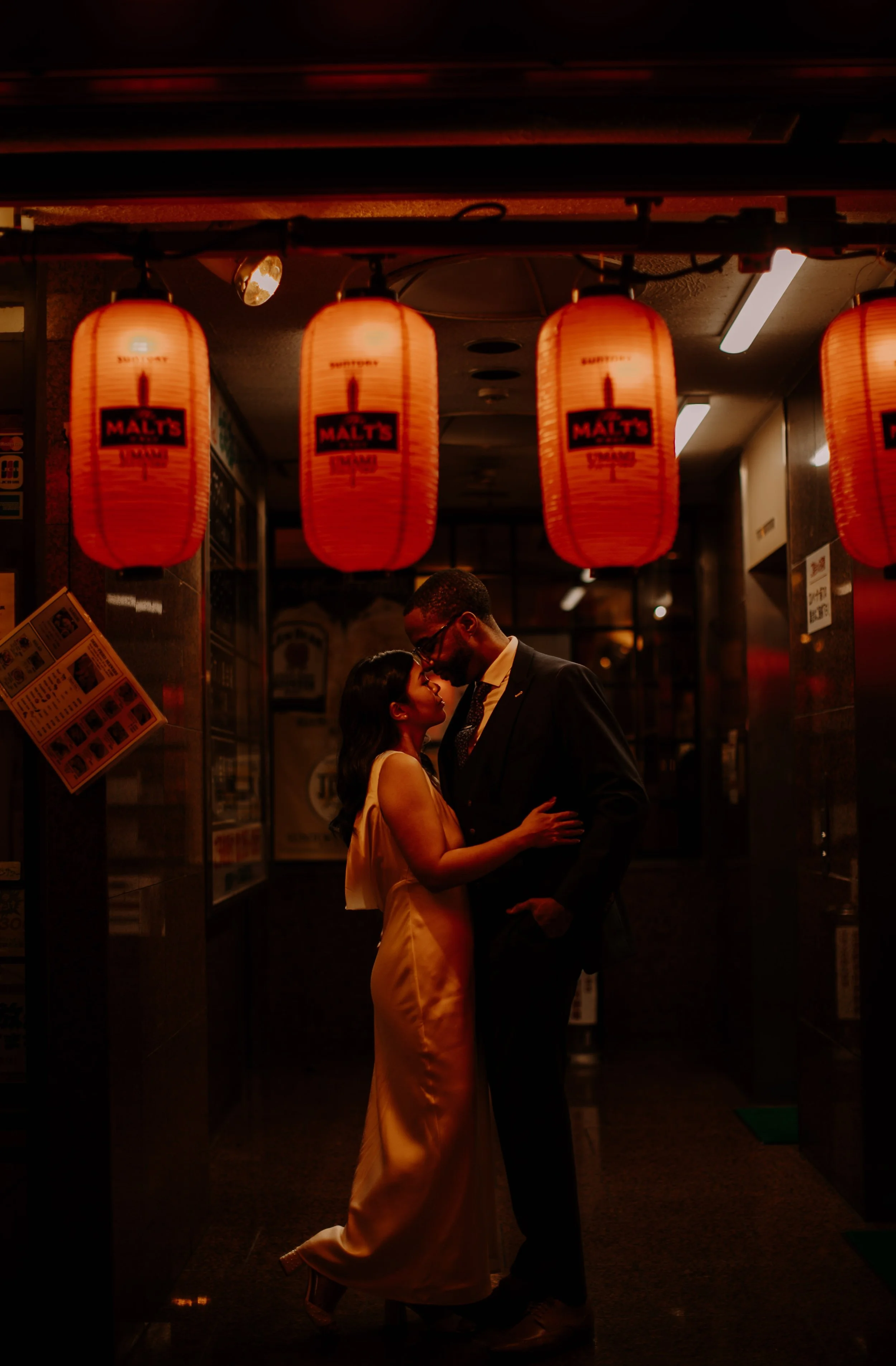 Couple sharing a kiss beneath glowing red lanterns in a Tokyo izakaya alley during their nighttime engagement photography session.
