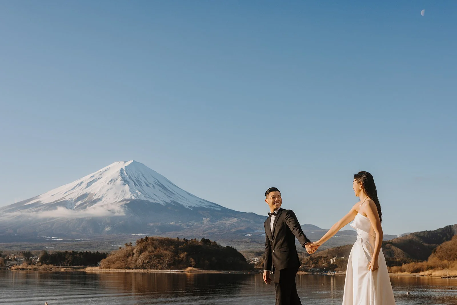 Couple holds hands and smiles at each other on the shoreline under a clear blue sky.