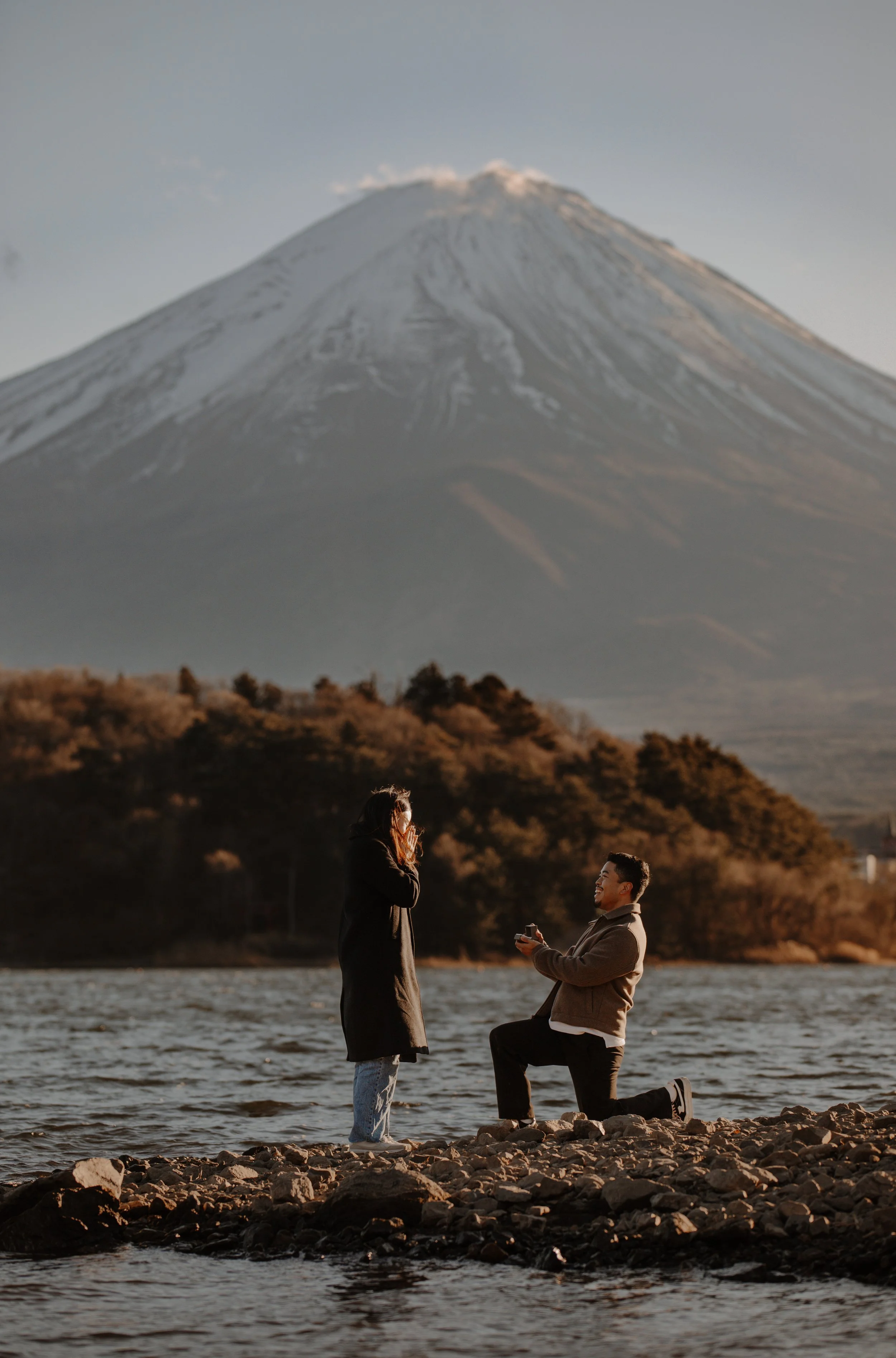 Man kneeling to propose beside the lake as his partner covers her face in emotion, Mount Fuji in the distance