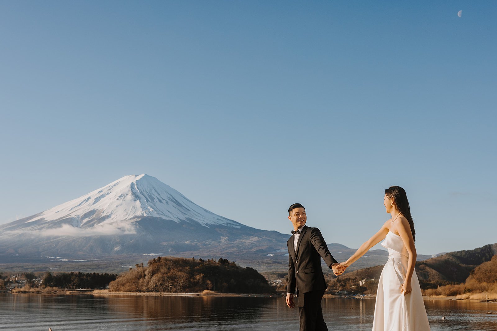 Couple holds hands and smiles at each other on the shoreline under a clear blue sky with Mt.Fuji in the back..