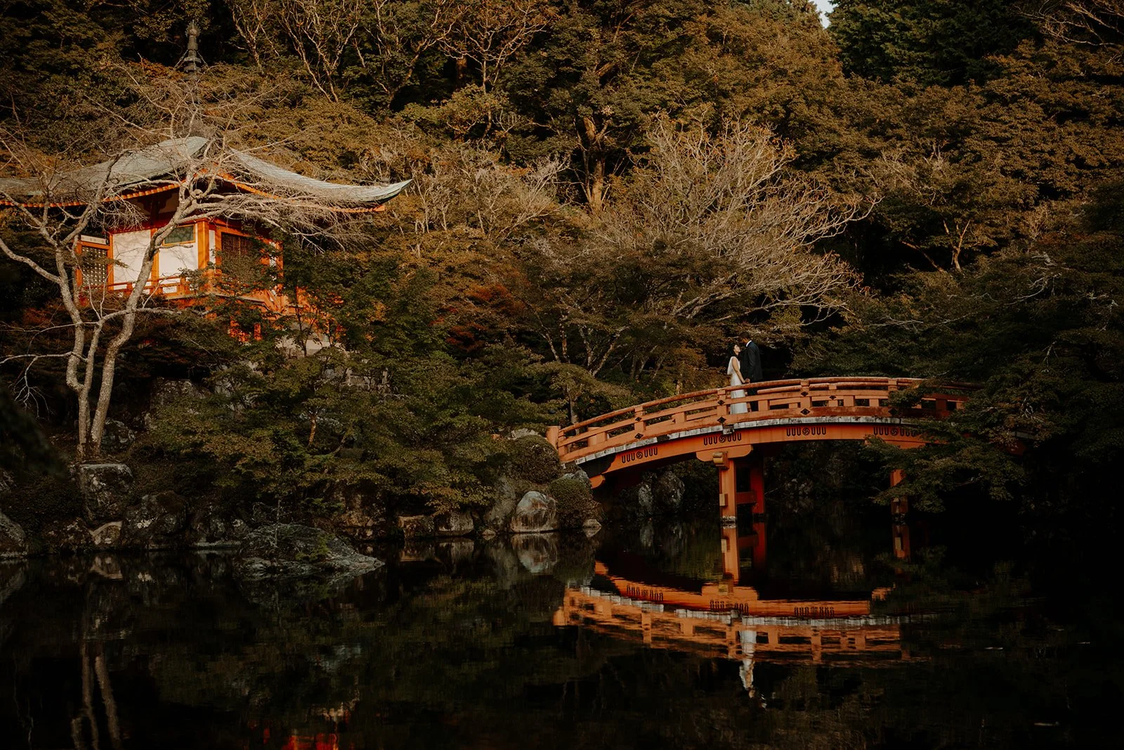 Couple holds hands on the arched red bridge below Daigo-ji's Bentendo hall, reflected in the pond.