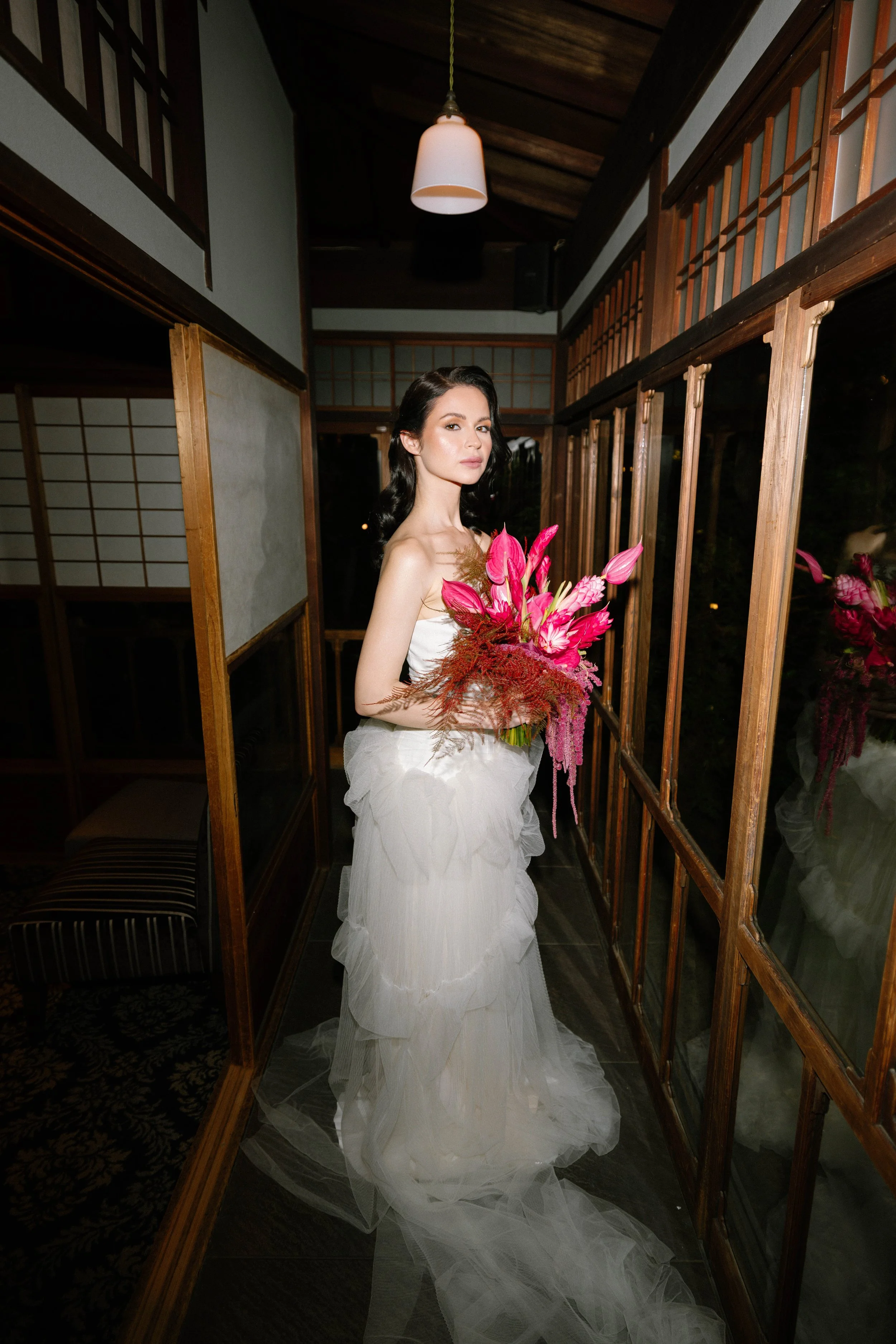 Bride alone in a dim traditional Japanese wooden corridor, holding a bouquet beneath a hanging lantern.
