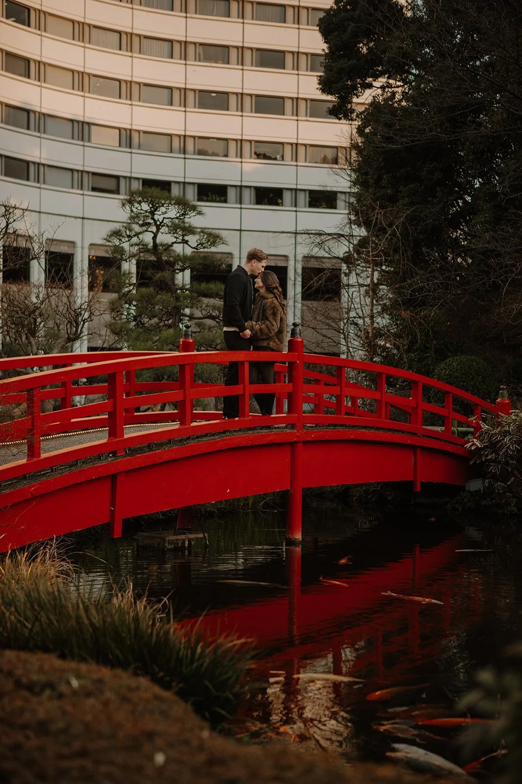Engaged couple embracing on a red garden bridge with a Tokyo hotel skyline reflected in the pond behind them.