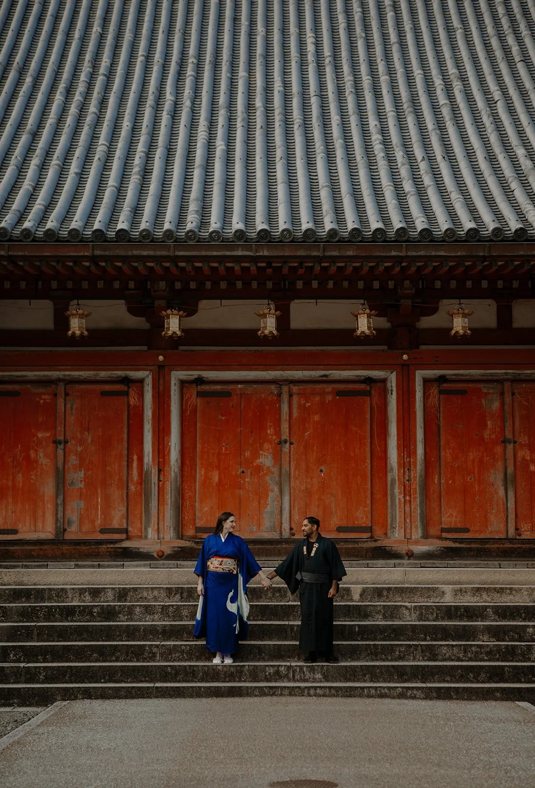 Couple in blue and black kimono holding hands on temple steps in front of a red gate in Kyoto, Japan