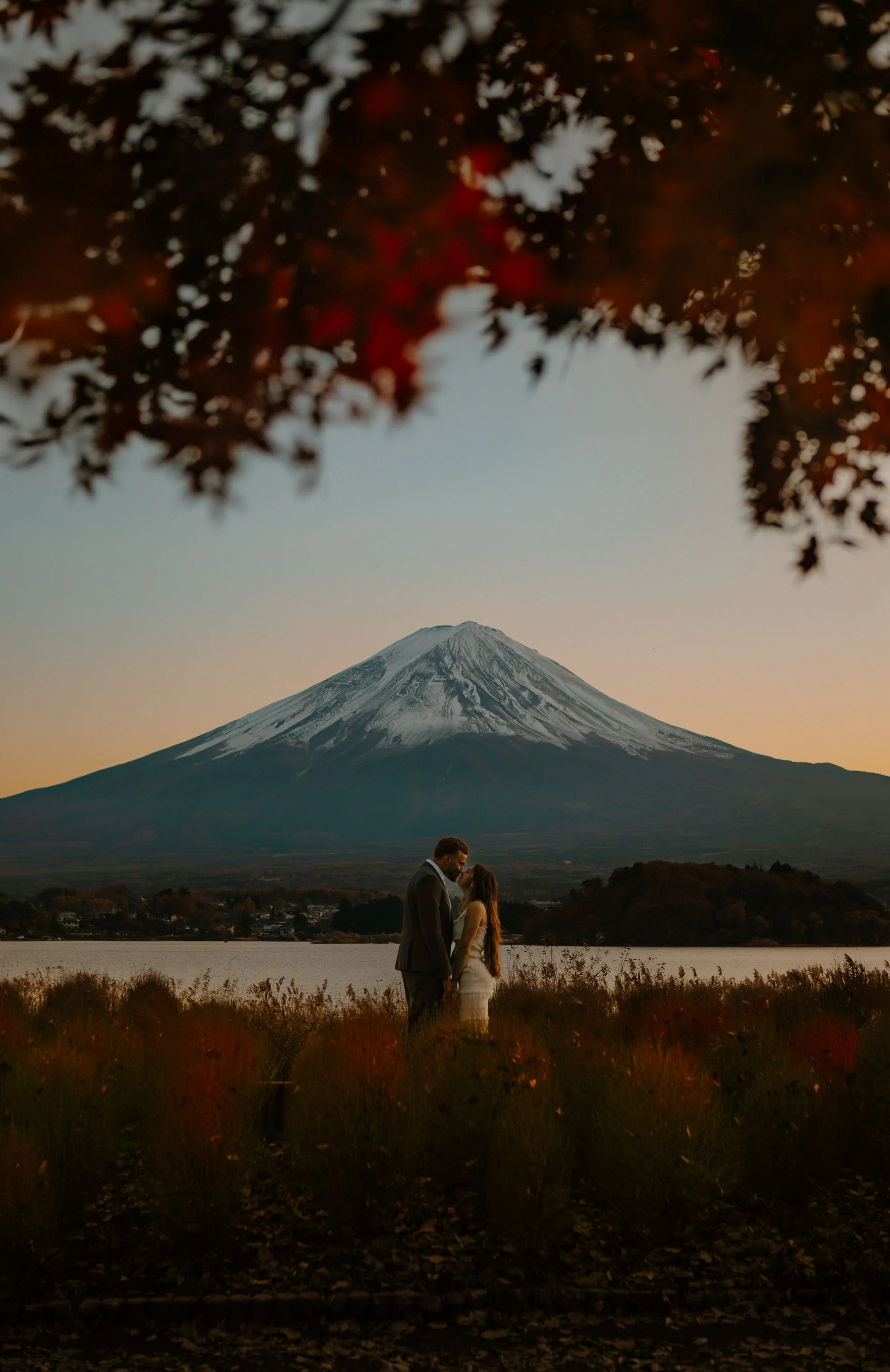 Couple kissing beneath a canopy of red autumn leaves with Mount Fuji and the lake glowing at sunset