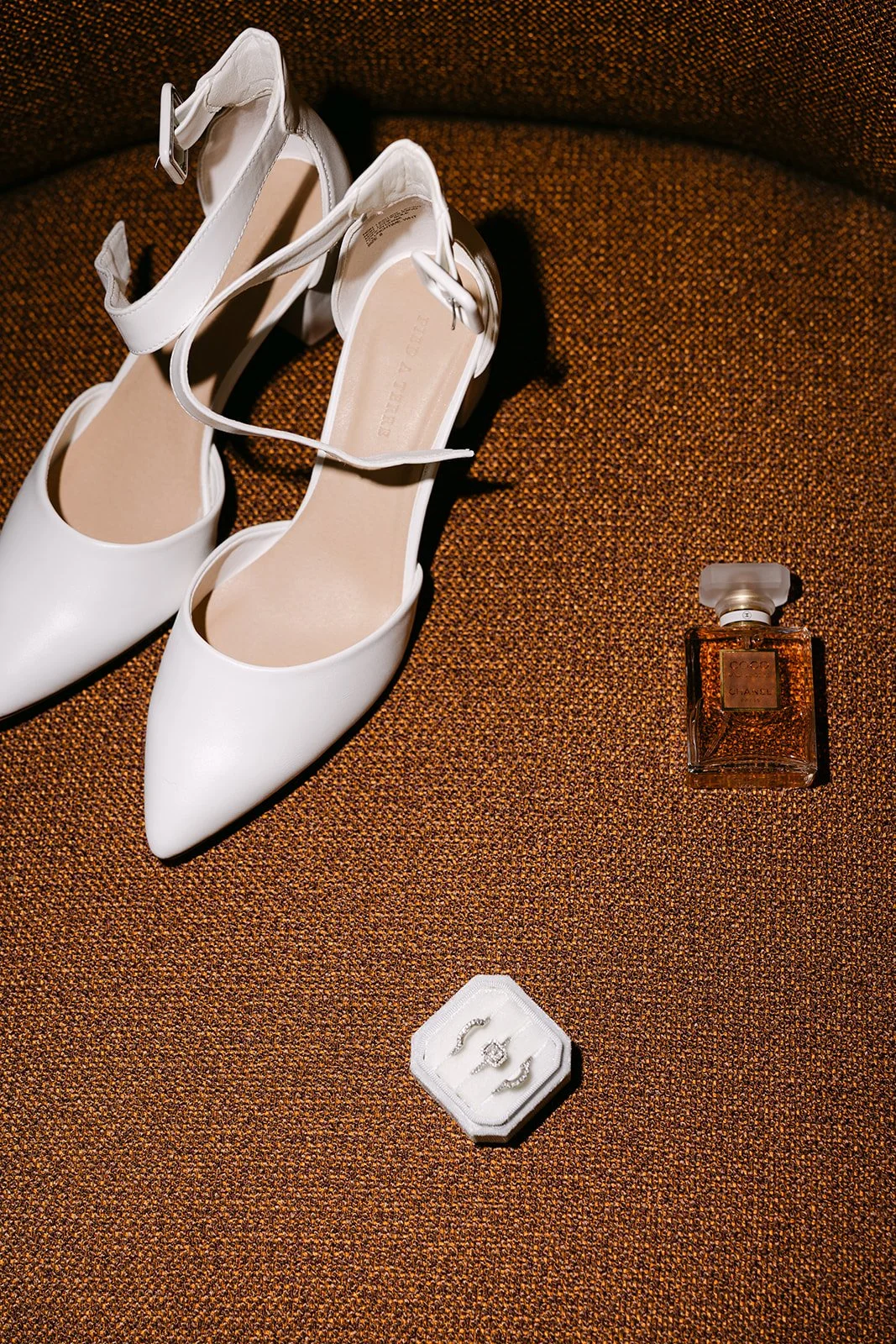 Bride's white heels, perfume, and engagement ring arranged on a textured chair indoors.