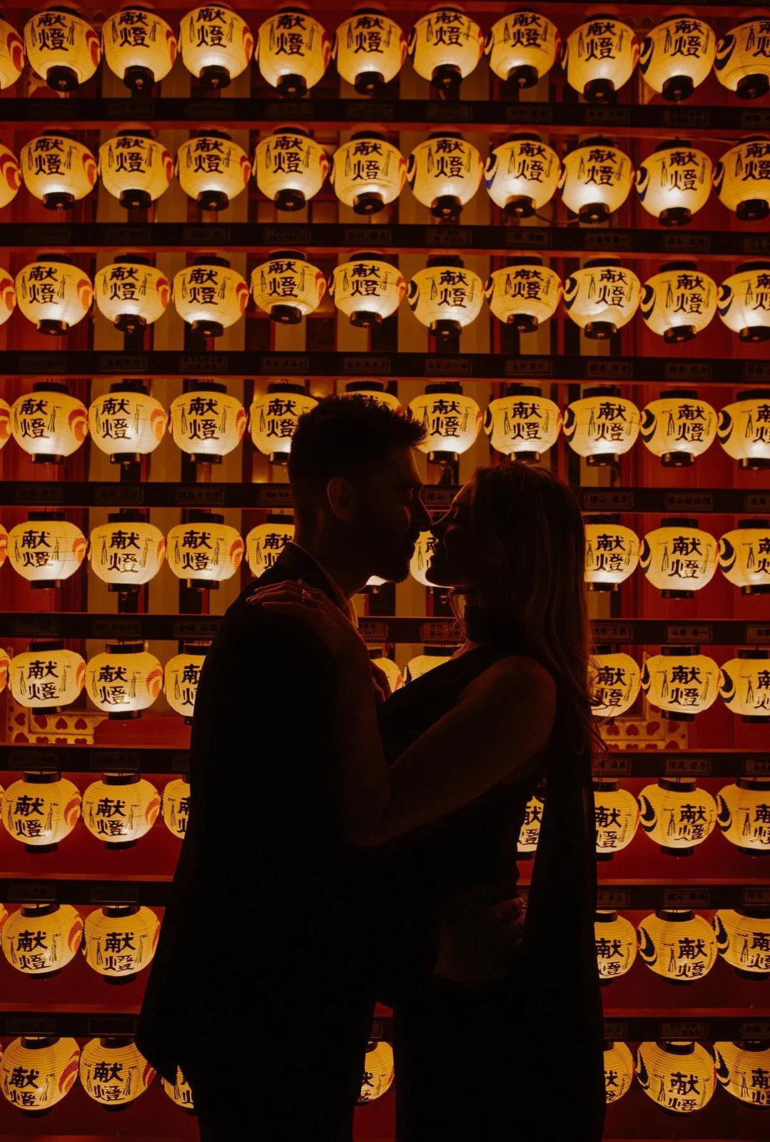 Silhouetted couple embracing in front of a glowing wall of paper lanterns at a Japanese shrine.