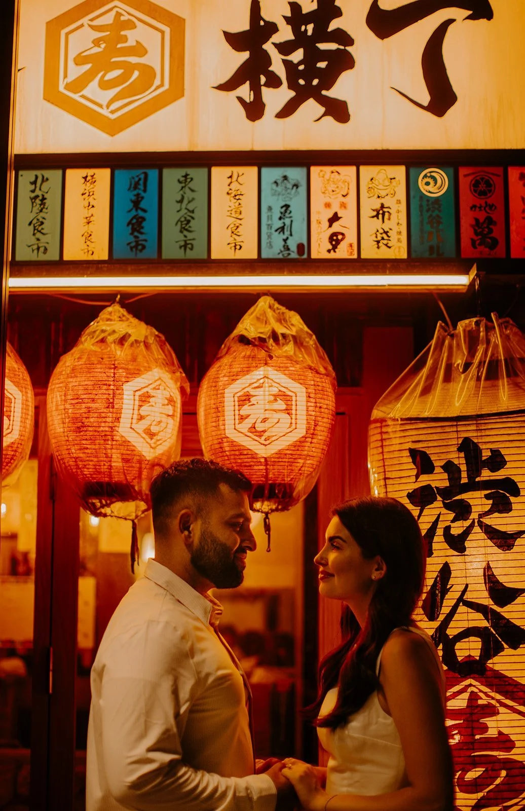 Couple holding hands under a lantern-lit food stall sign in a Tokyo izakaya alley at night