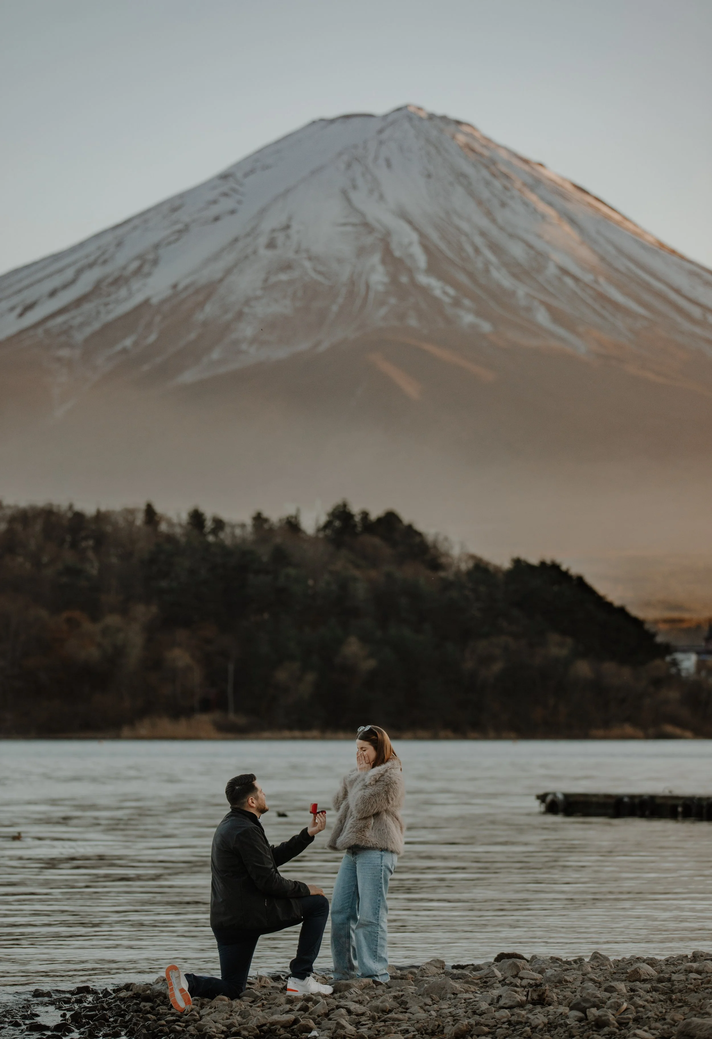 Man kneeling to propose on a rocky lakeshore as woman reacts with emotion, snow-dusted Mount Fuji in the mist