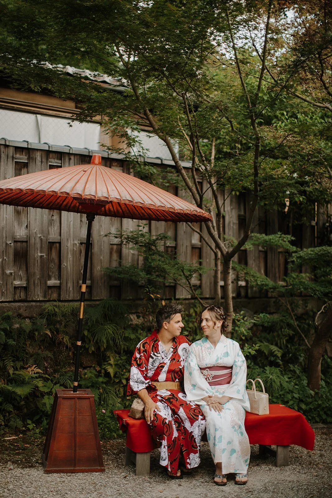 Couple in traditional kimono sitting together under a red paper parasol in a Kyoto garden