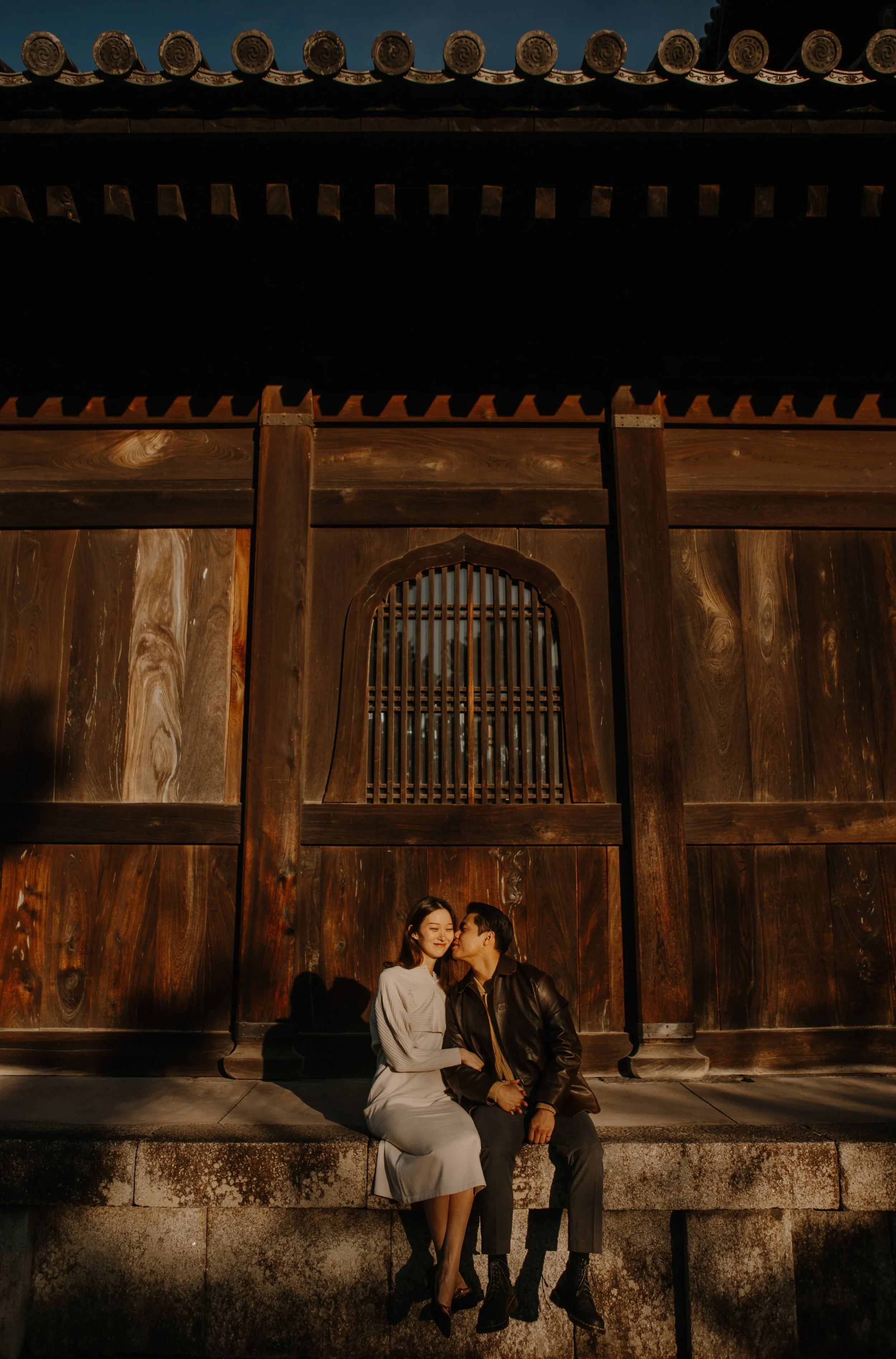 Couple sitting together on a stone wall beneath the towering wooden eaves of a temple gate, warm sunset light on their faces.