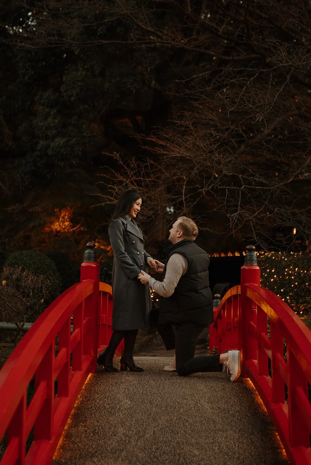 Man kneeling to propose to his partner on a red bridge in a Tokyo hotel garden at dusk