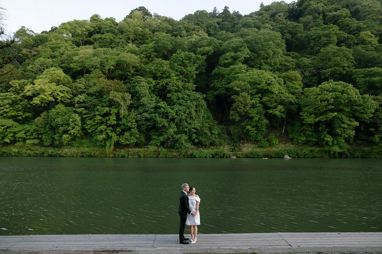 Couple embraces on a wooden dock beside a wide green river in Tokyo.