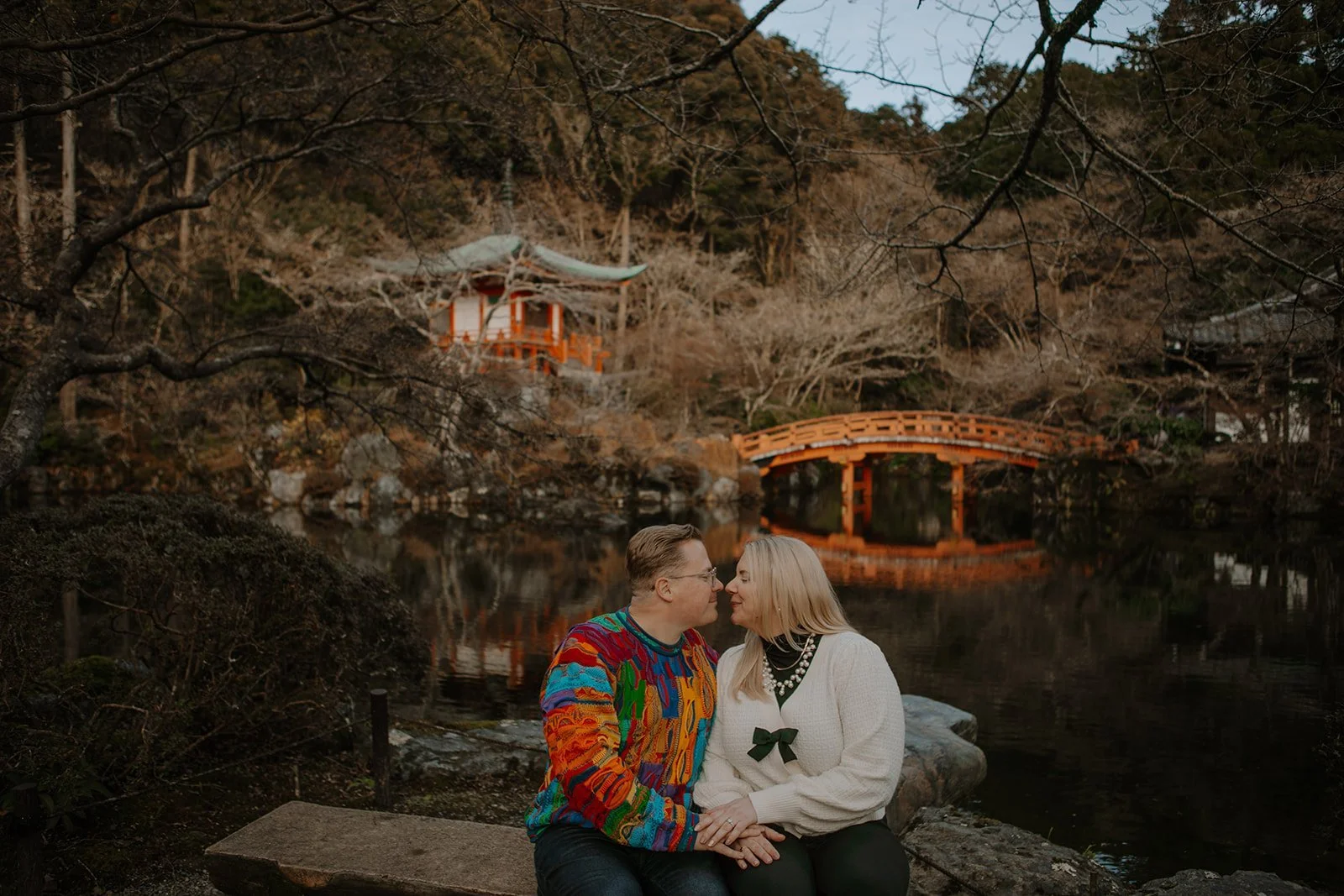 Client testimonial photo of a couple embracing beside a reflecting pond and red bridge at a Kyoto temple garden