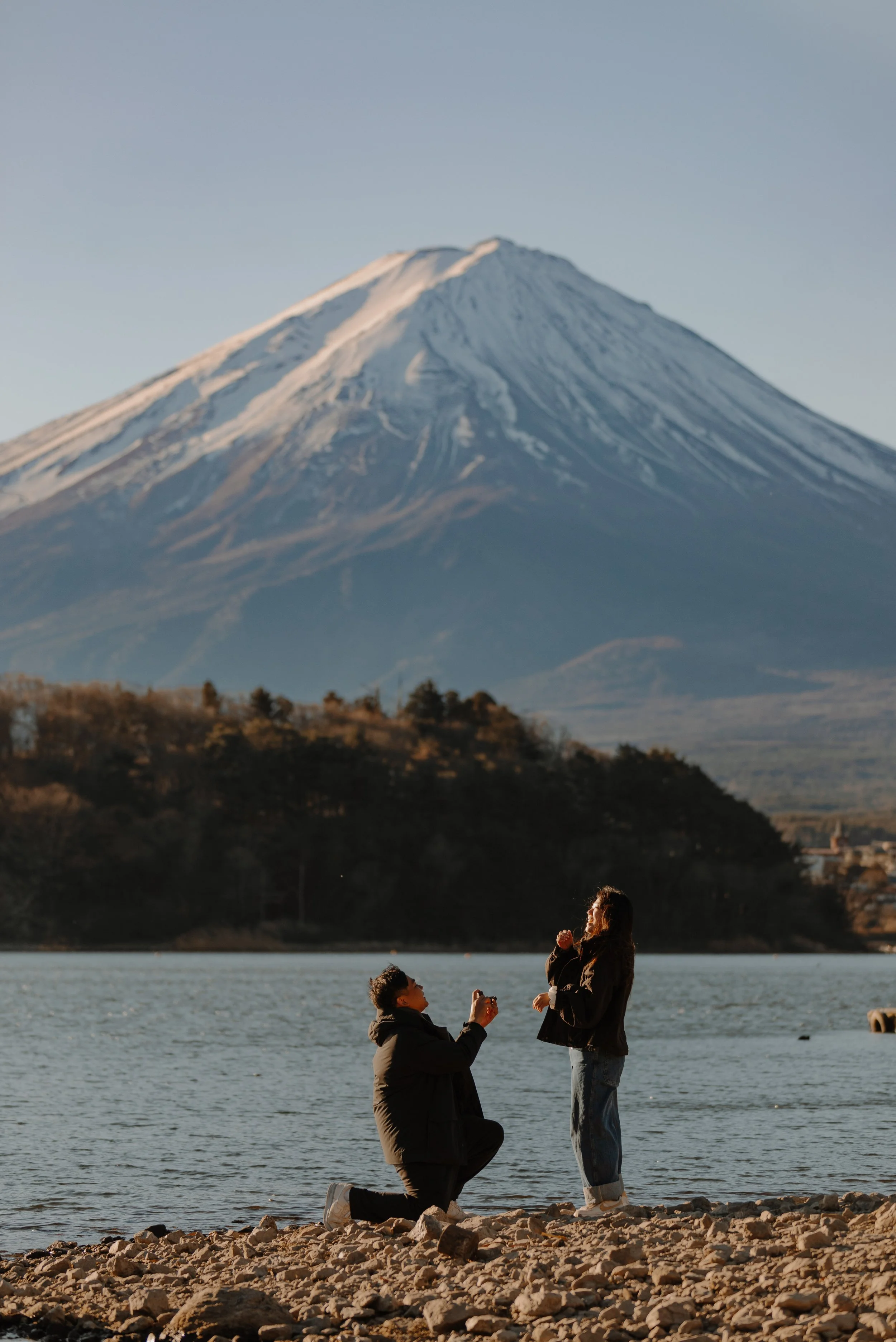 Man kneeling to propose beside Lake Kawaguchiko at sunset as his partner reacts with joy, Mount Fuji behind