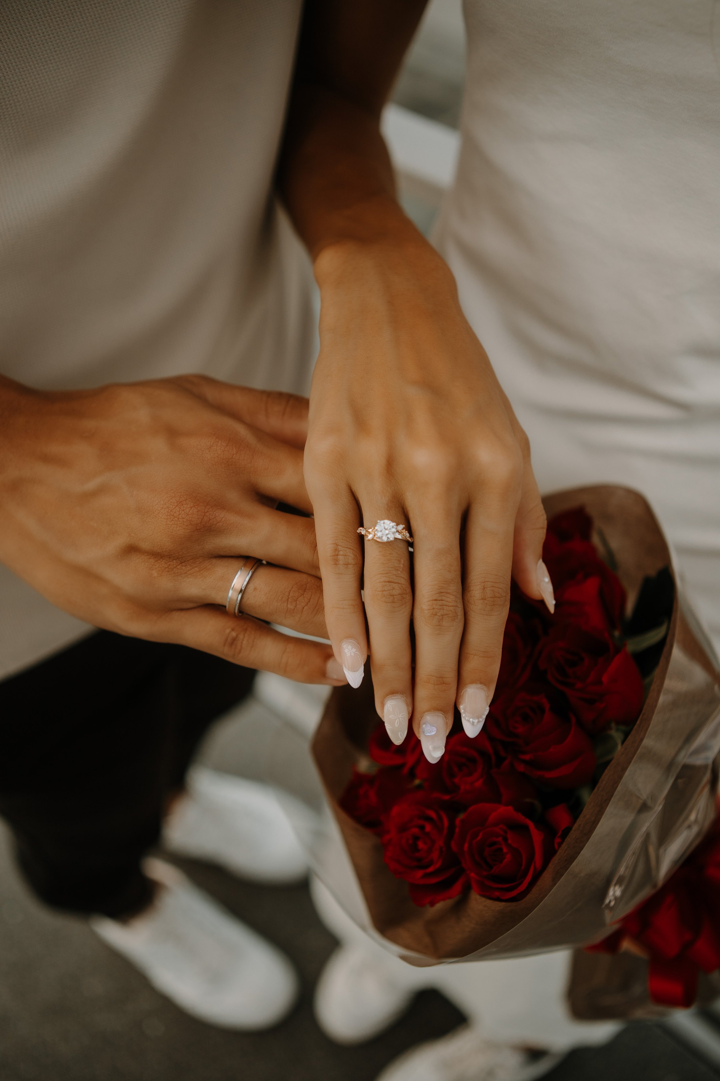 Close-up of hands with engagement and wedding rings resting on a bouquet of red roses