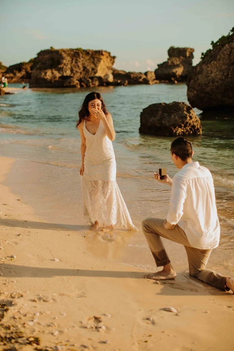 Man kneeling to propose on an Okinawa beach as his partner covers her mouth in surprise, rock formations behind